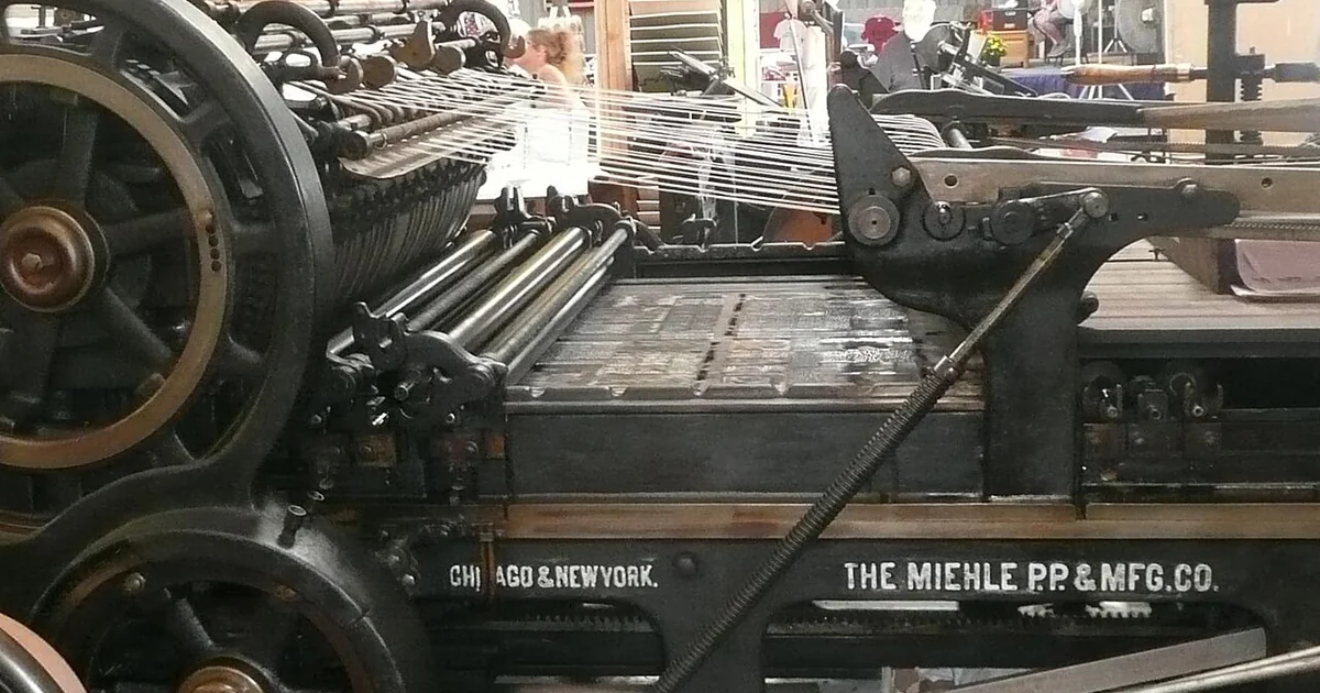 The bed of metal type passing under the inking rollers of a Miehle Model 00 flat-bed cylinder printing press. The print roller is on the left; the press operator, in the upper left, loads pages to be printed, while the webbing above the bed