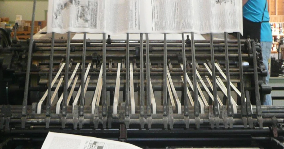 A Miehle Model 00 flat-bed cylinder printing press in operation, transferring a freshly printed page to the output stack. The press operator, behind the page on the right, is loading the next page to be printed. This press, now at the print