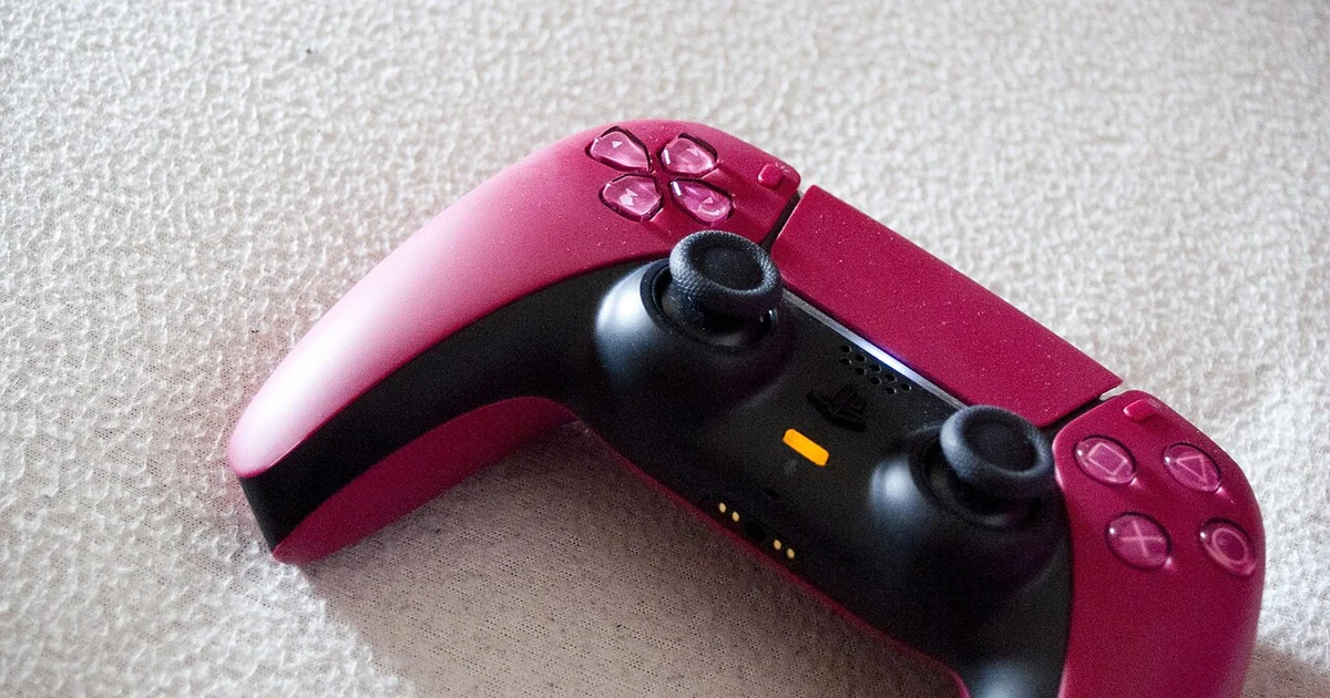 A red and black PlayStation controller rests on a light, textured fabric surface. The controller features two black analog sticks, a directional pad on the left side, and the characteristic symbol buttons (triangle, circle, cross, and squar
